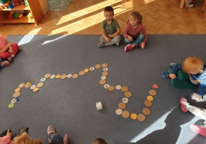Dzieci grają w dużą gre planszową na dywanie. (6 zadanie z akcji Code Week)